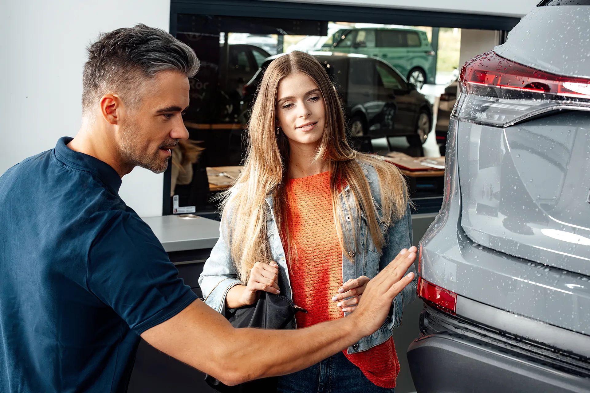Ein Unfall Spezialist im Gespräch mit einer Kundin zeigt auf die Delle eines grauen Fahrzeugs.
