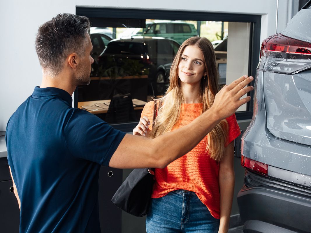 Ein Unfall Spezialist im Gespräch mit einer Kundin zeigt auf das Heck eines grauen Fahrzeugs.